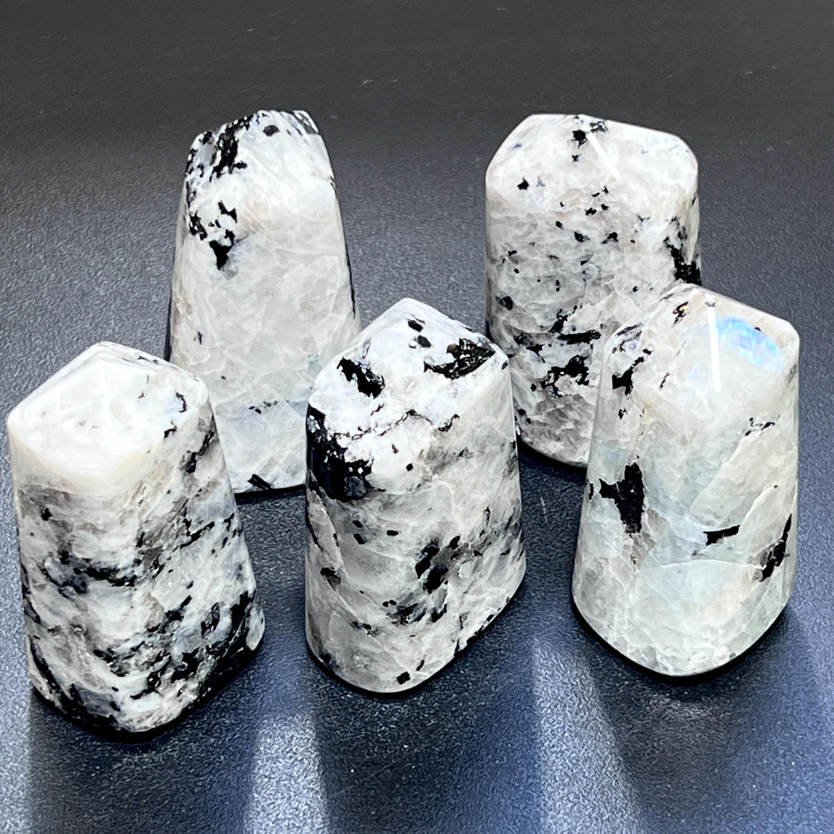 Five standing rainbow moonstone point towers showing white and gray patterns, dark inclusions, and a blue flash on a dark surface.

