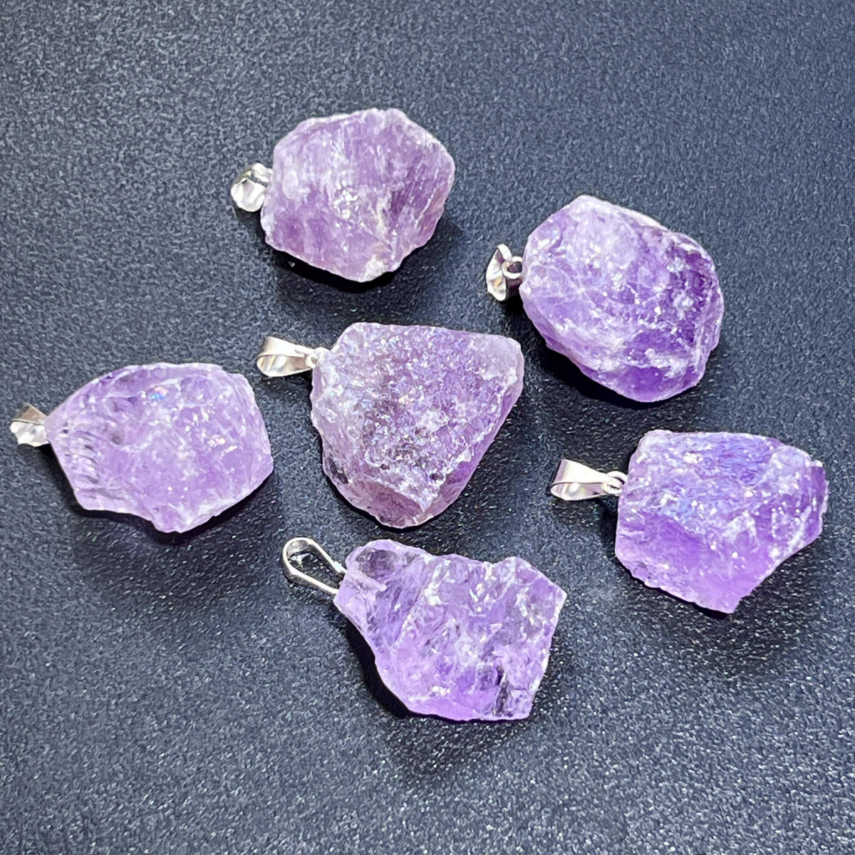 Six irregular raw amethyst pendants showing textured purple surfaces and silver-tone hanging bails.
