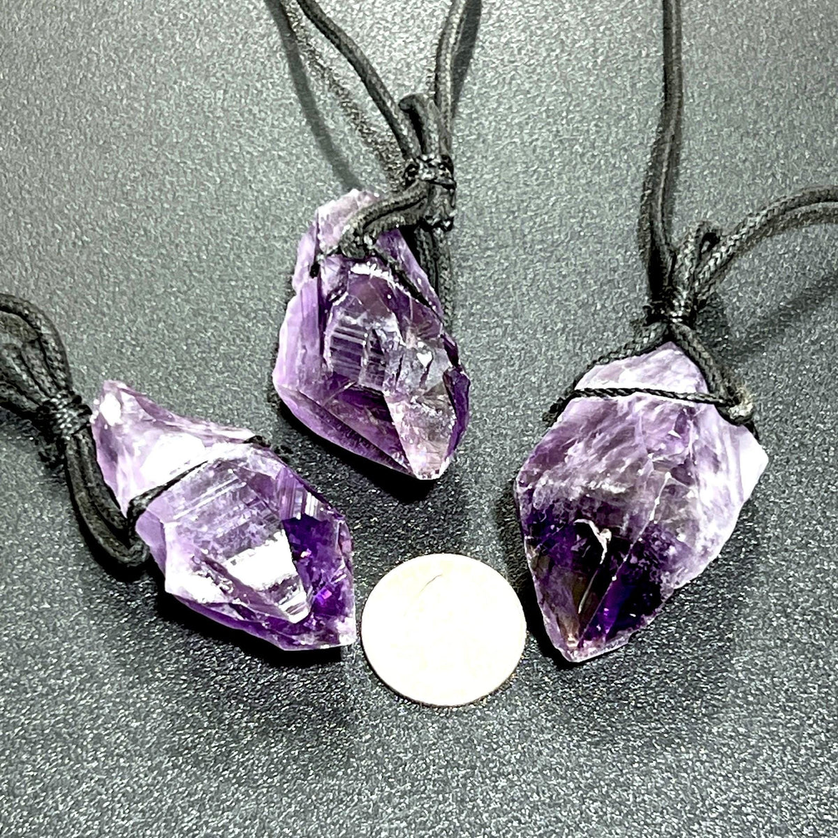 Three raw amethyst point necklaces with black cords beside a coin for size comparison.