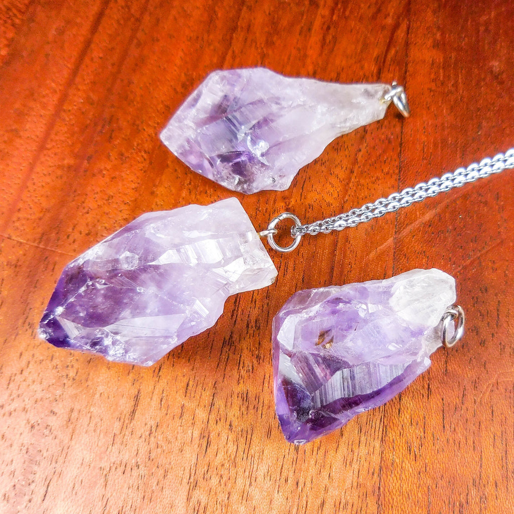 Close-up of three raw amethyst point pendants with silver-tone rings and a silver-colored chain on wood.
