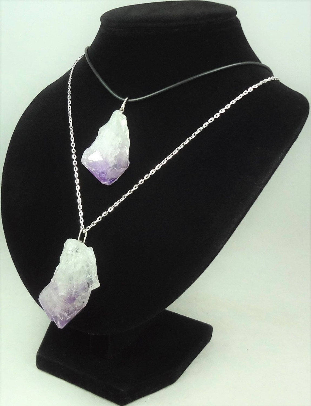 Two raw amethyst point pendants displayed on a black necklace bust, one on a black cord and one on a silver-colored chain.
