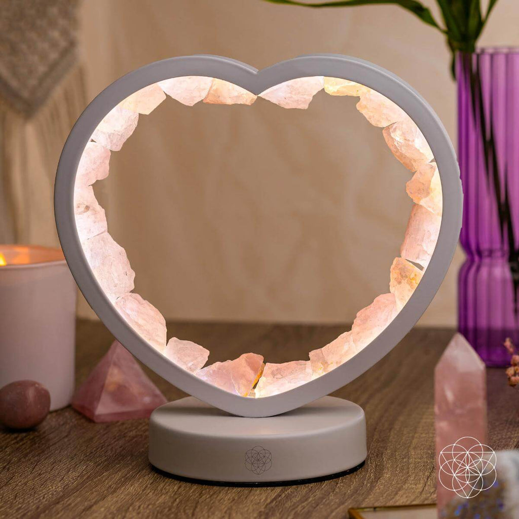 Glowing rose quartz heart lamp on a wooden tabletop beside a purple vase, candle, and pink crystal points.
