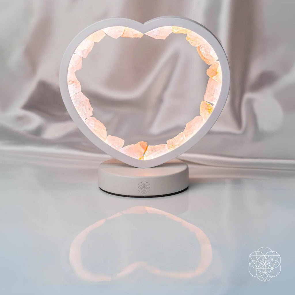 Lit rose quartz heart lamp reflected in a glossy white surface against draped pale fabric.

