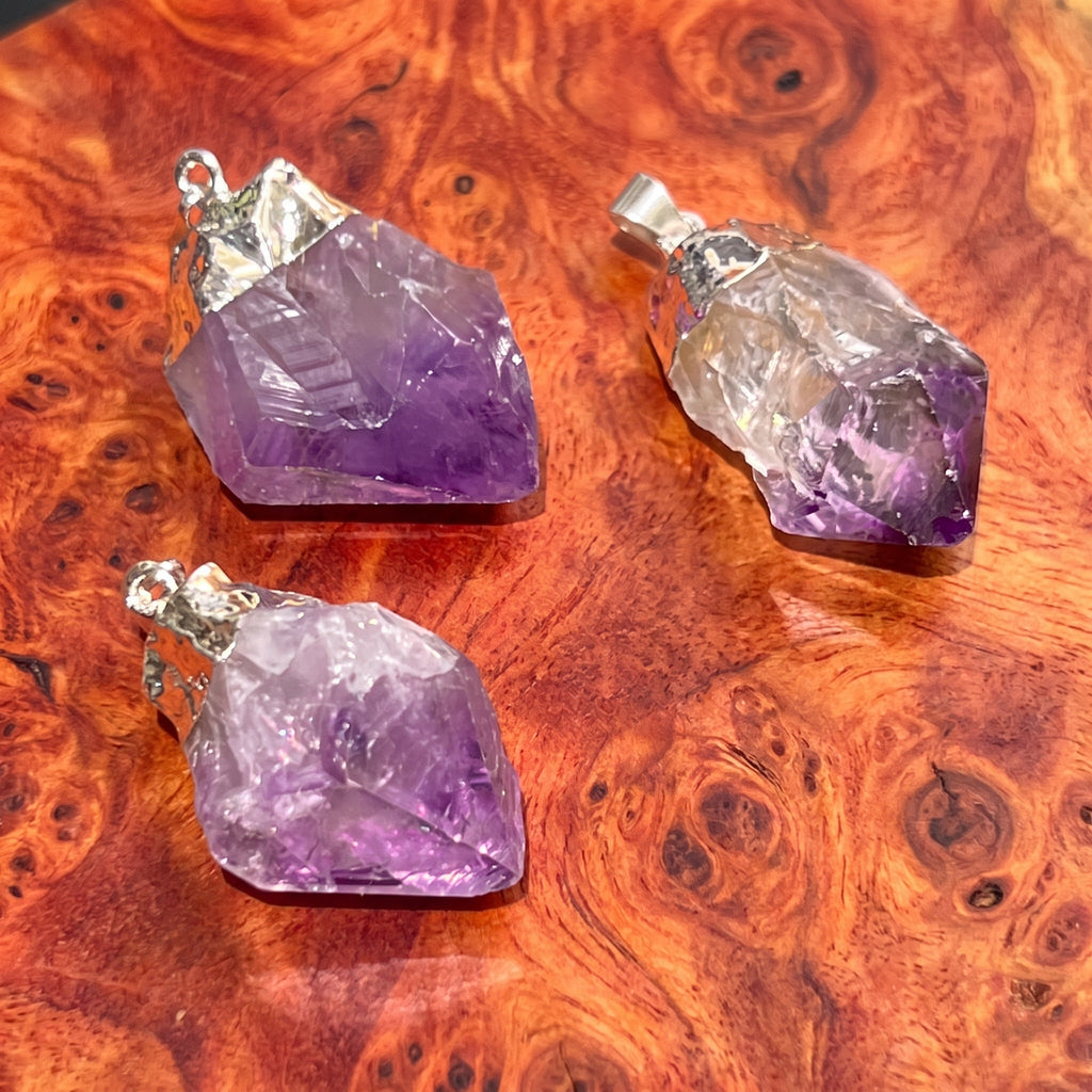 Three rough purple amethyst pendants with silver-plated fittings on reddish figured wood.
