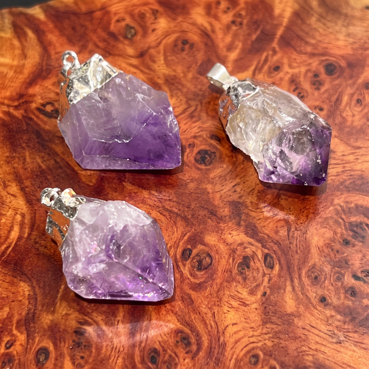 Three raw amethyst crystal pendants with silver bail loops, displayed on a richly textured brown burl wood surface.
