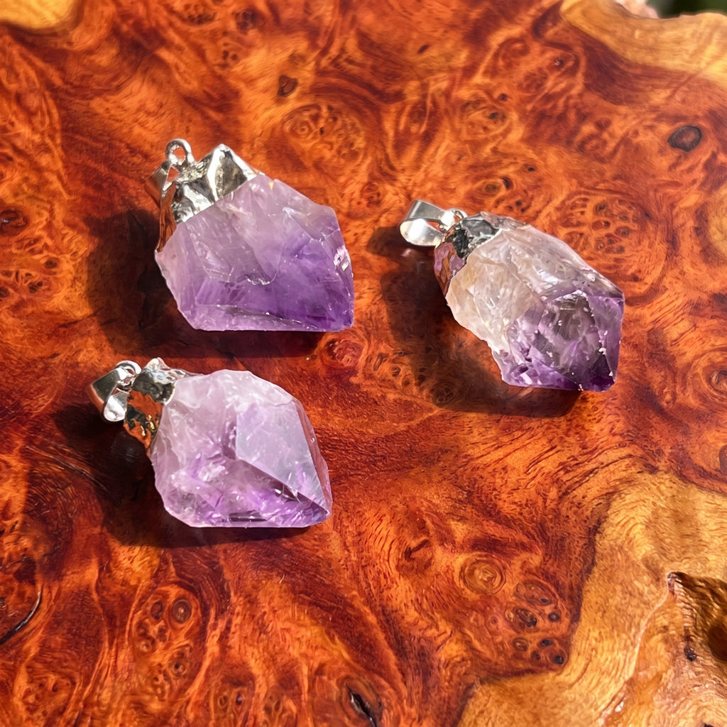Three raw amethyst crystal pendants with silver bail loops, displayed on a richly textured, wavy brown wood surface.