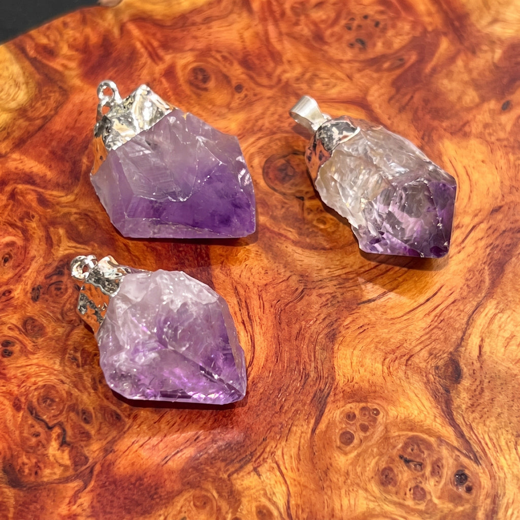 Three raw amethyst crystal pendants with silver bail loops, displayed on a richly textured, swirling brown wood surface.
