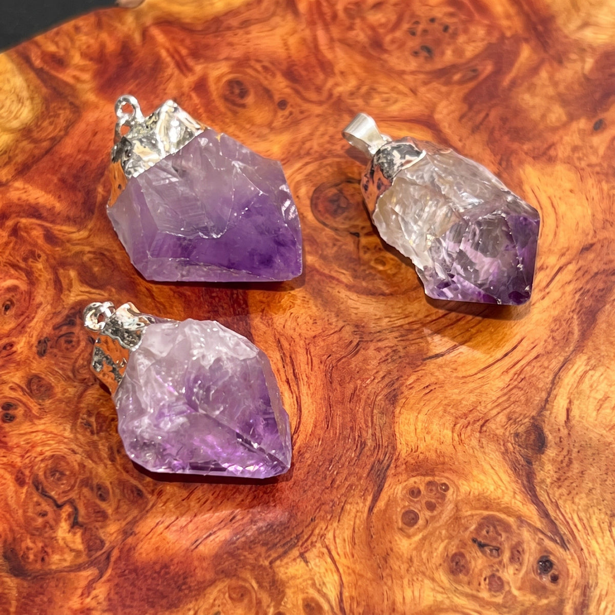 Three raw amethyst crystal pendants with silver bail loops, displayed on a richly textured, swirling brown wood surface.
