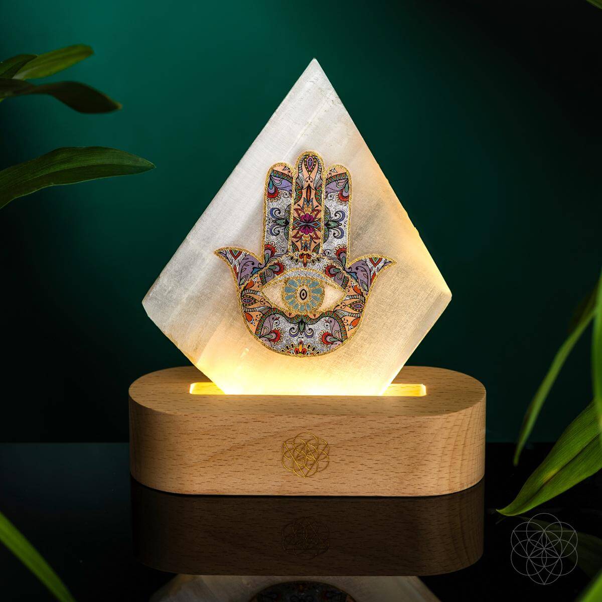 Illuminated selenite Hamsa hand lamp on a rounded wooden base against a teal background with green leaves
