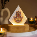 Glowing selenite panel lamp with a colorful Hamsa hand and eye motif on a wooden base beside candles
