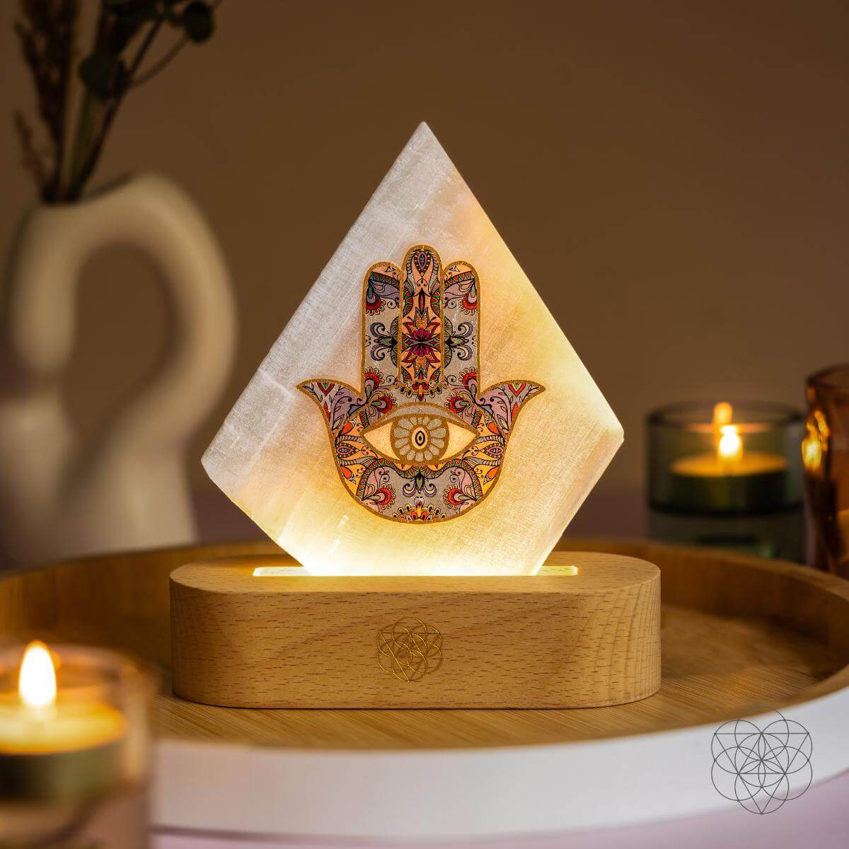 Glowing selenite panel lamp with a colorful Hamsa hand and eye motif on a wooden base beside candles
