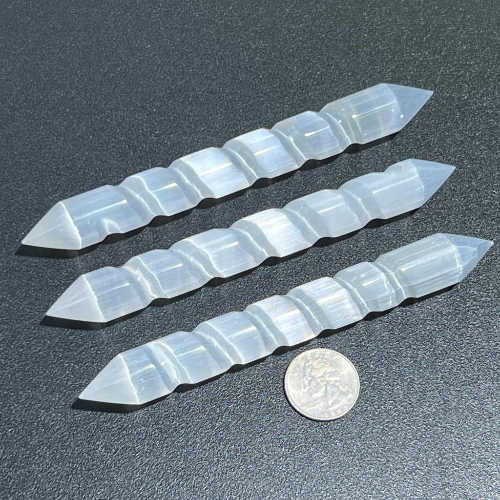 Three double-pointed selenite spiral wands beside a quarter for size comparison on a dark surface.