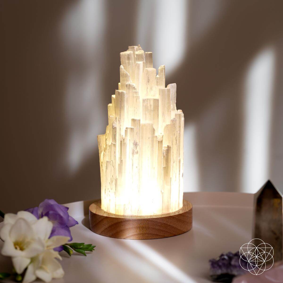 Illuminated selenite tower lamp on a walnut base beside white and purple flowers and decorative crystals.
