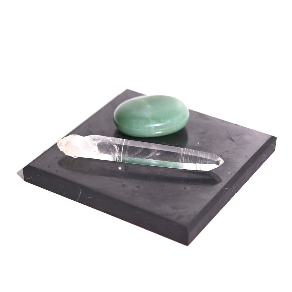 Black square shungite plate styled with a clear quartz point and a polished green stone on a white background.
