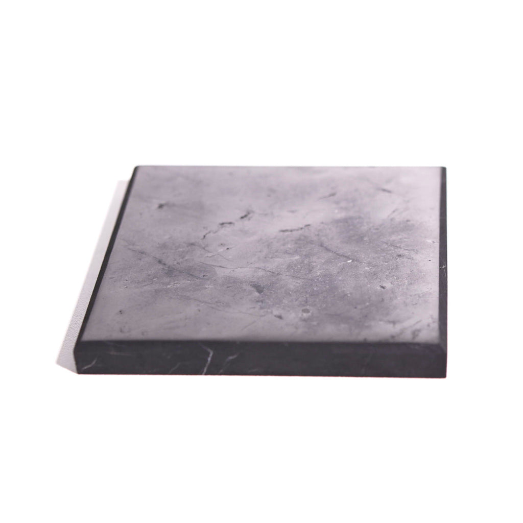 Low angled view of a square shungite plate showing a gray-black surface and thick dark front edge.
