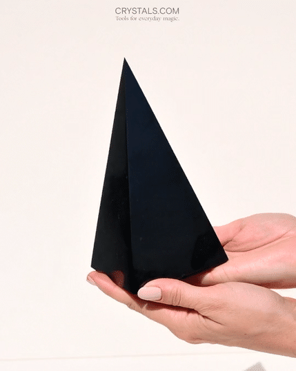 Two hands supporting a tall polished black shungite pyramid against a pale background.
