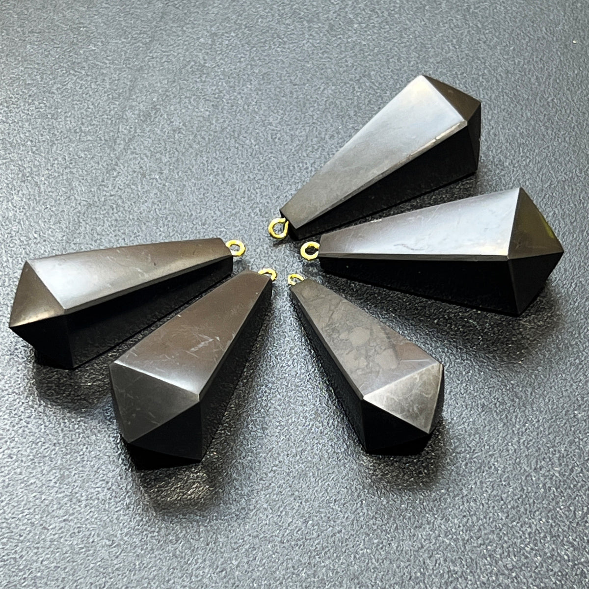 Five faceted shungite pendulum pendants with subtle surface markings arranged on a dark textured background.
