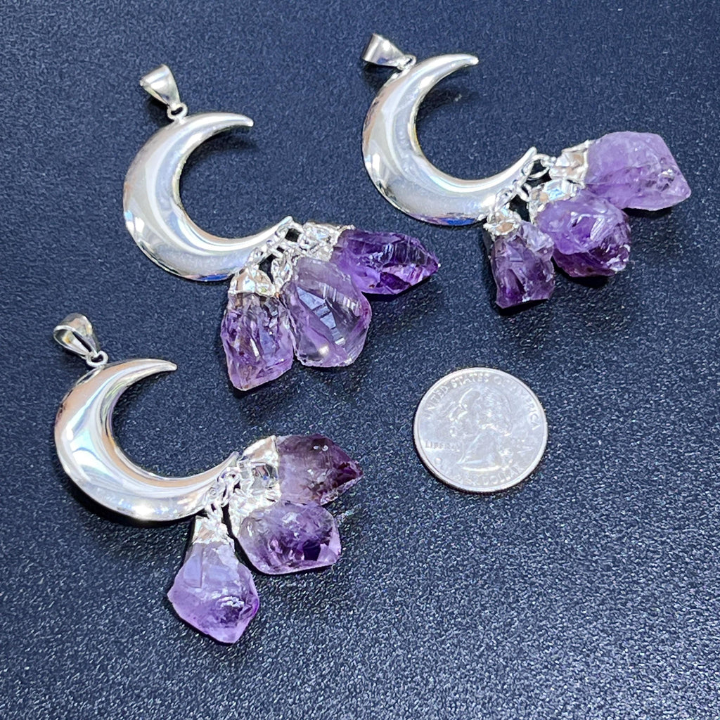 Three silver-plated crescent moon pendants with dangling purple amethyst crystals beside a quarter for scale.
