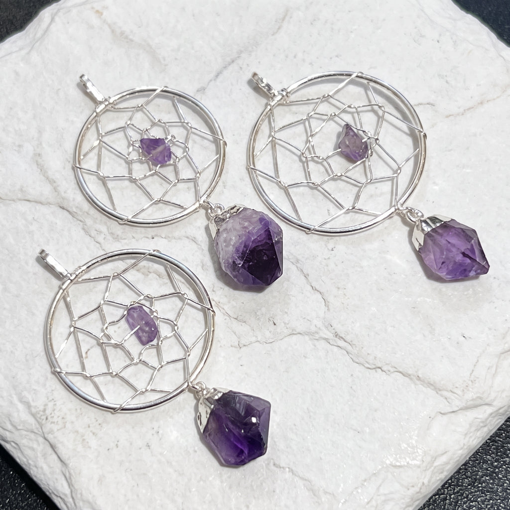 Three silver-plated dreamcatcher pendants with purple amethyst accents displayed on a round white stone surface