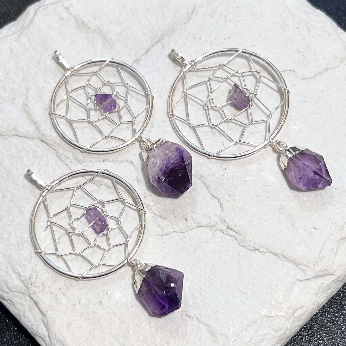 Three silver-plated dreamcatcher pendants with purple amethyst accents displayed on a round white stone surface