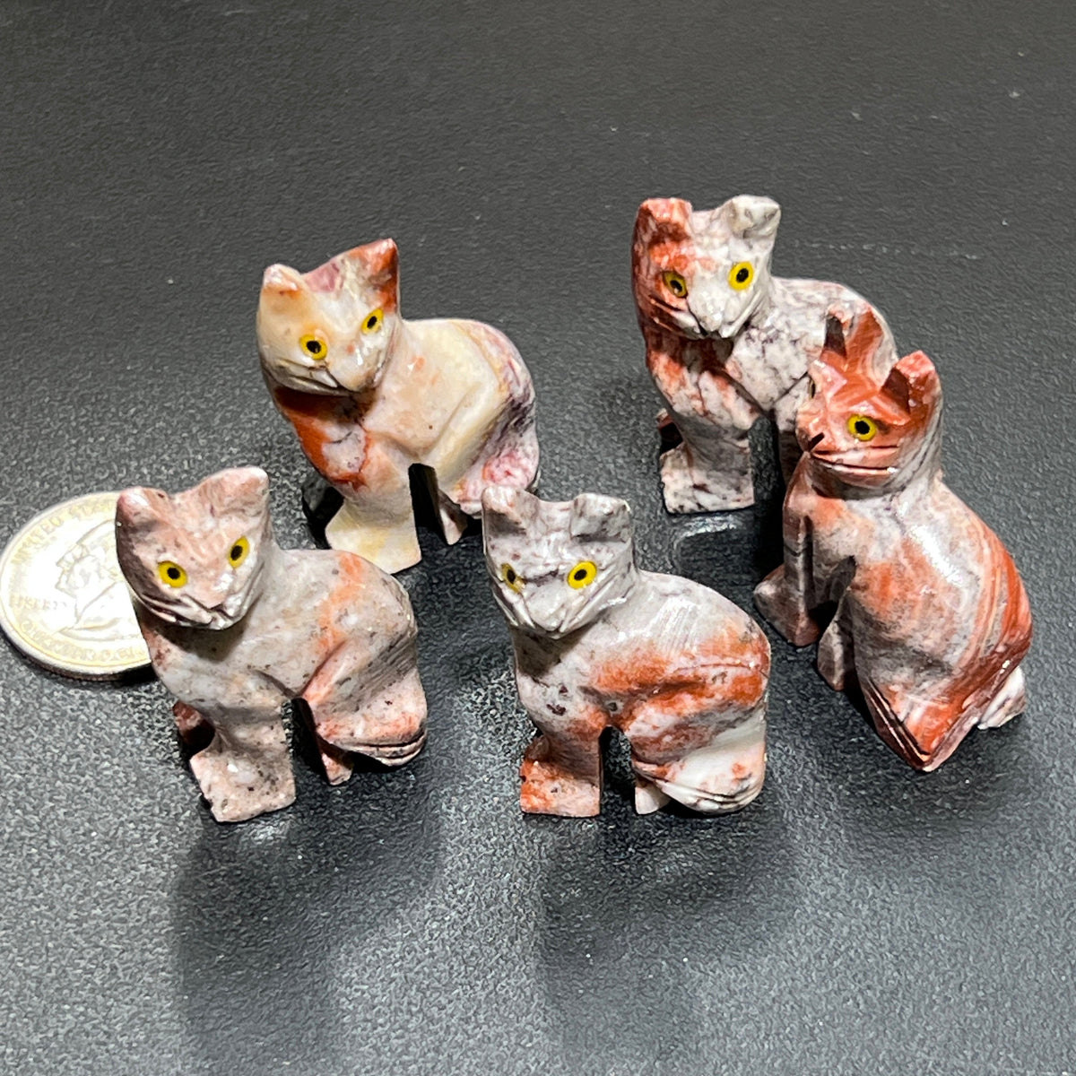 Five miniature soapstone cat carvings with yellow eyes and varied pink, cream and gray markings beside a coin