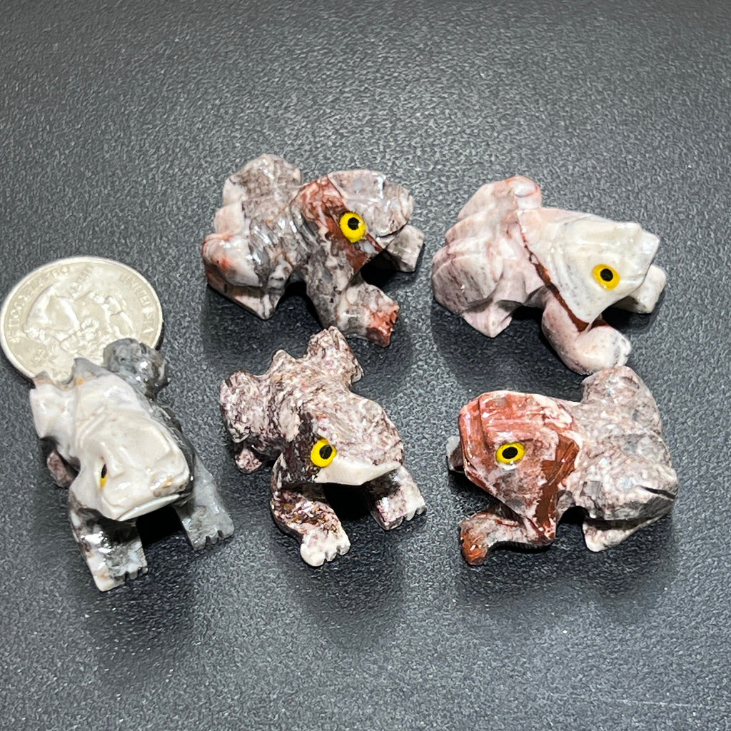 Five miniature soapstone frog carvings with yellow eyes and varied pink, cream and gray patterns beside a coin