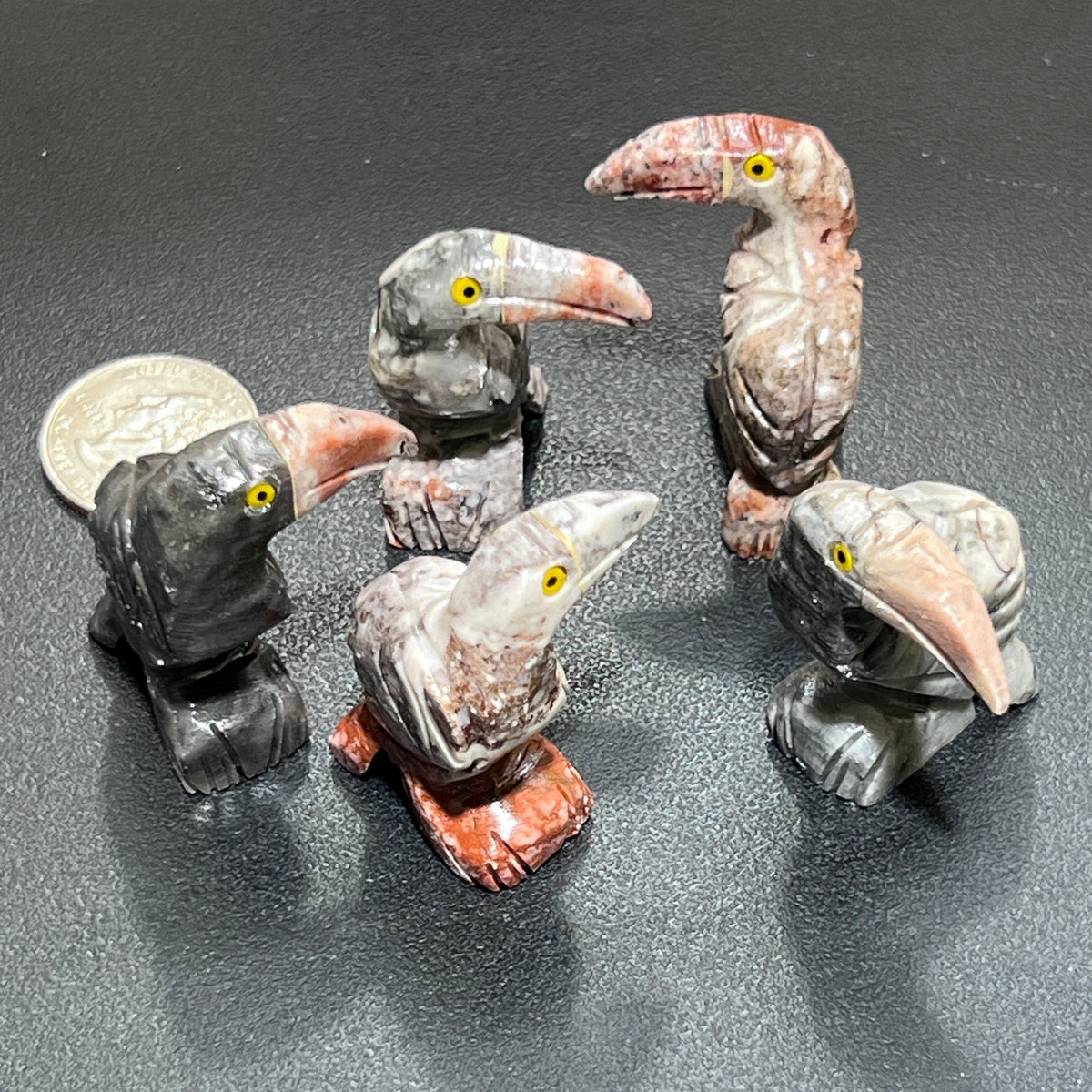 Five soapstone toucan carvings with long beaks and yellow eyes in gray, cream and rust tones beside a coin