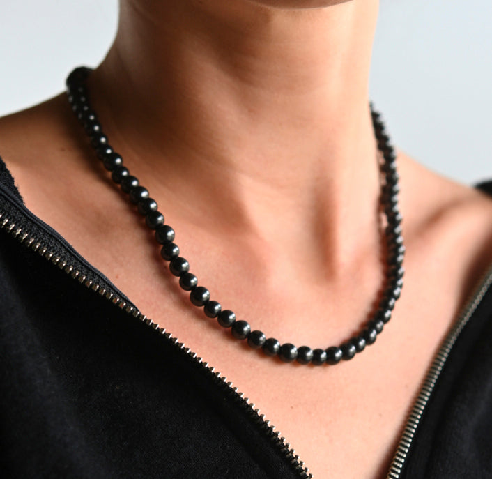Close-up of round black shungite beads worn as a necklace above a black open neckline.
