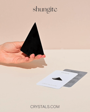 Hand holding a tall black shungite pyramid beside collector cards on a pale surface.
