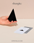 Hand holding a tall black shungite pyramid beside collector cards on a pale surface.
