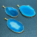 Three teal agate slice pendants with banded patterns, gold-plated edges, and hanging bails on a dark surface.
