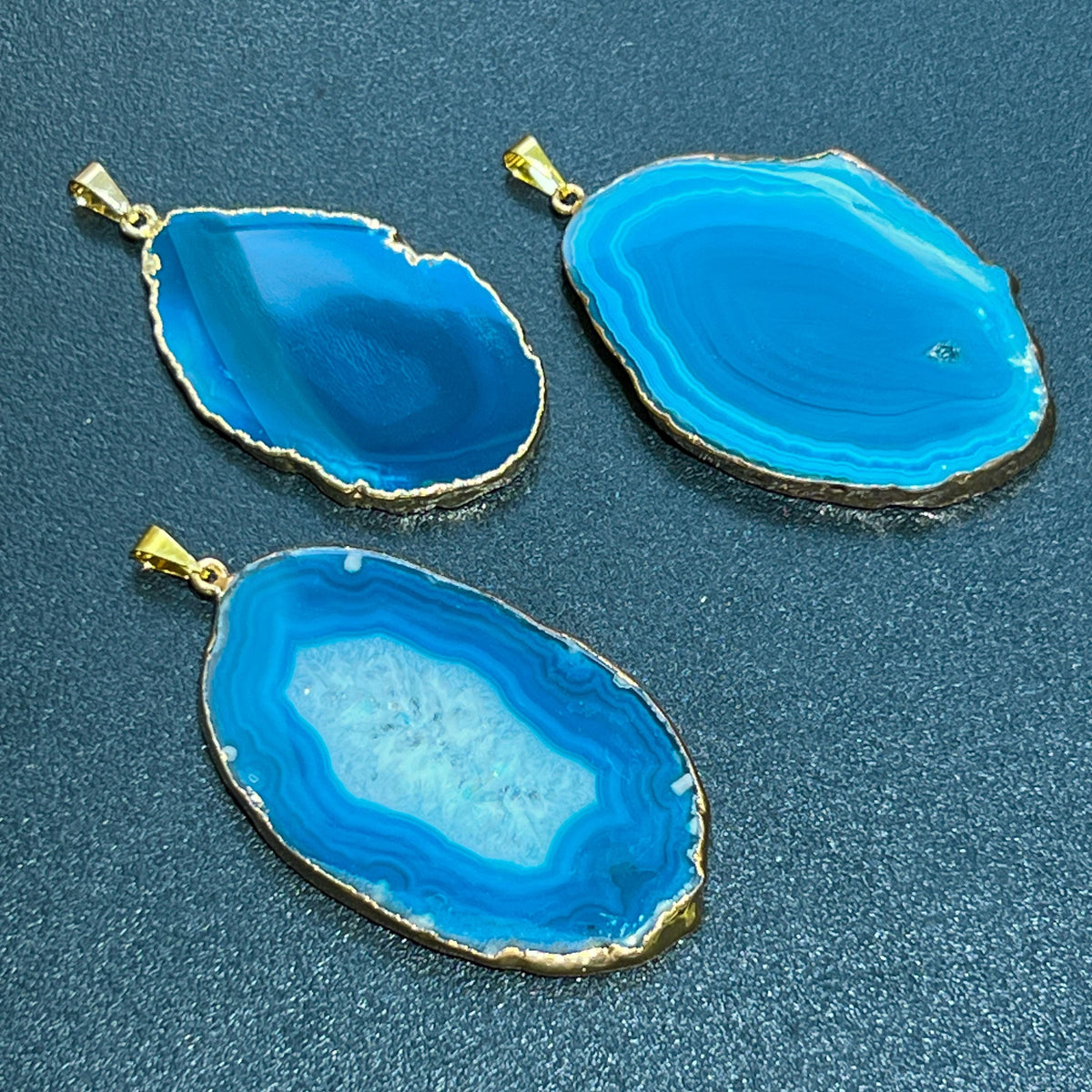Three teal agate slice pendants with banded patterns, gold-plated edges, and hanging bails on a dark surface.
