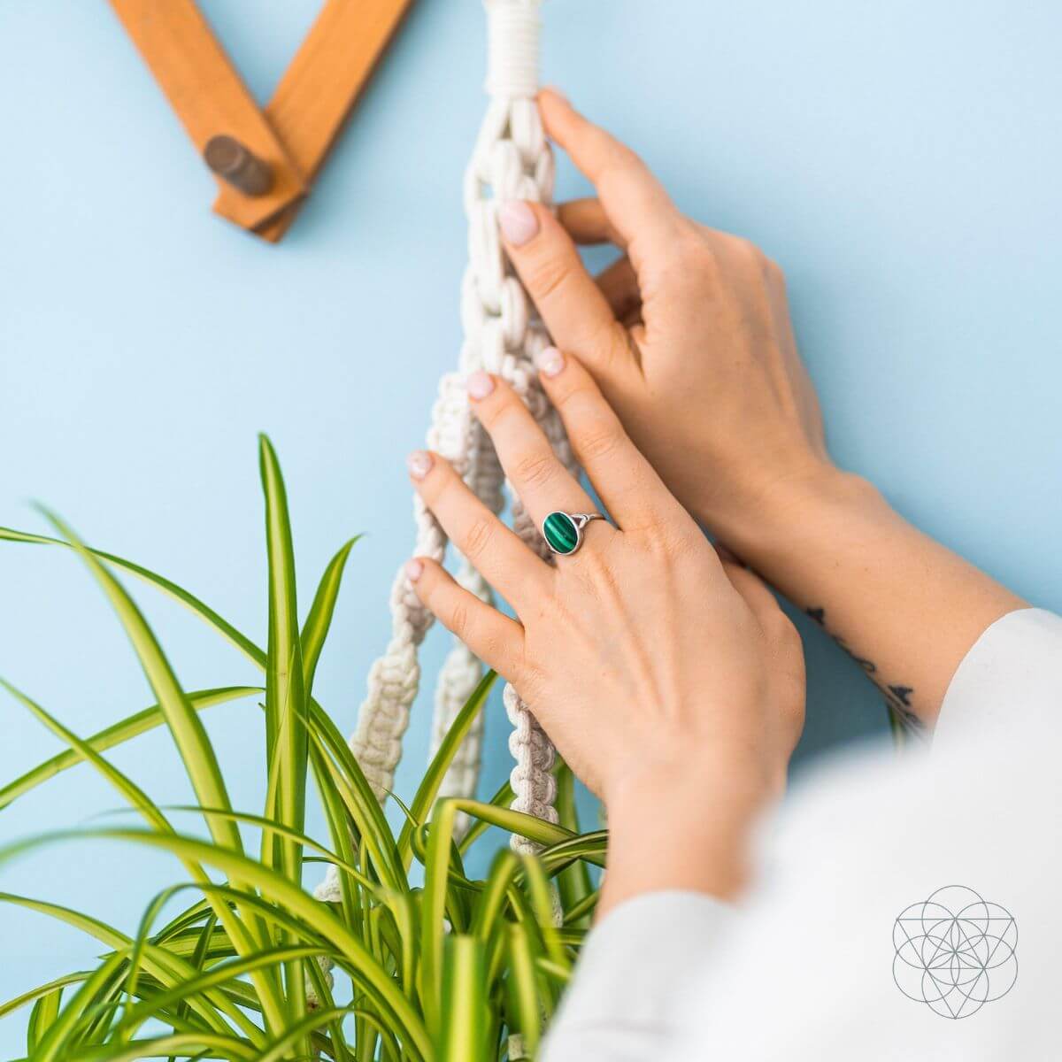 Close-up of an oval malachite ring worn on a hand beside a hanging plant against a blue wall.
