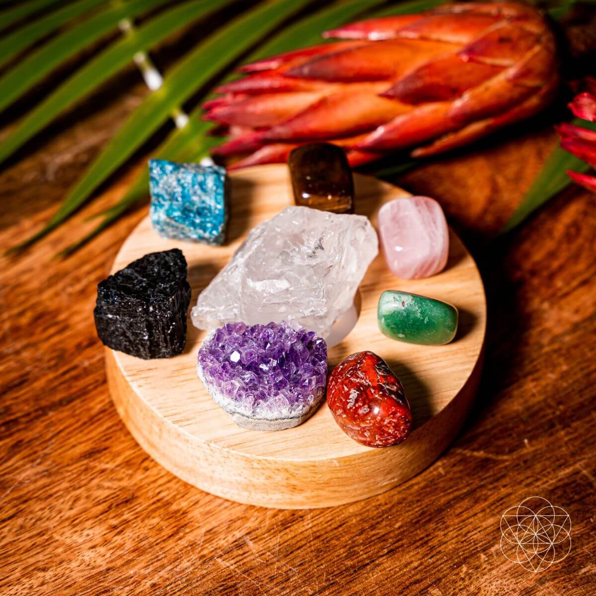 Mixed rough and polished crystals arranged around clear quartz on a round wooden lamp base beside a red flower.

