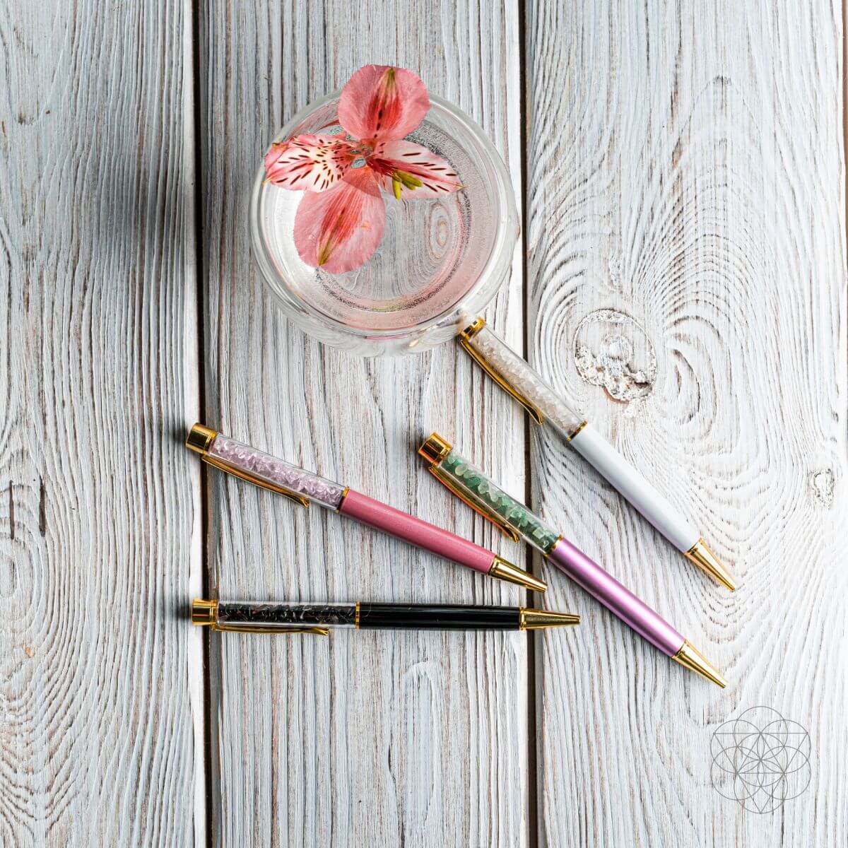 Four colorful crystal pens on whitewashed wood beside a glass bowl holding a pink flower.
