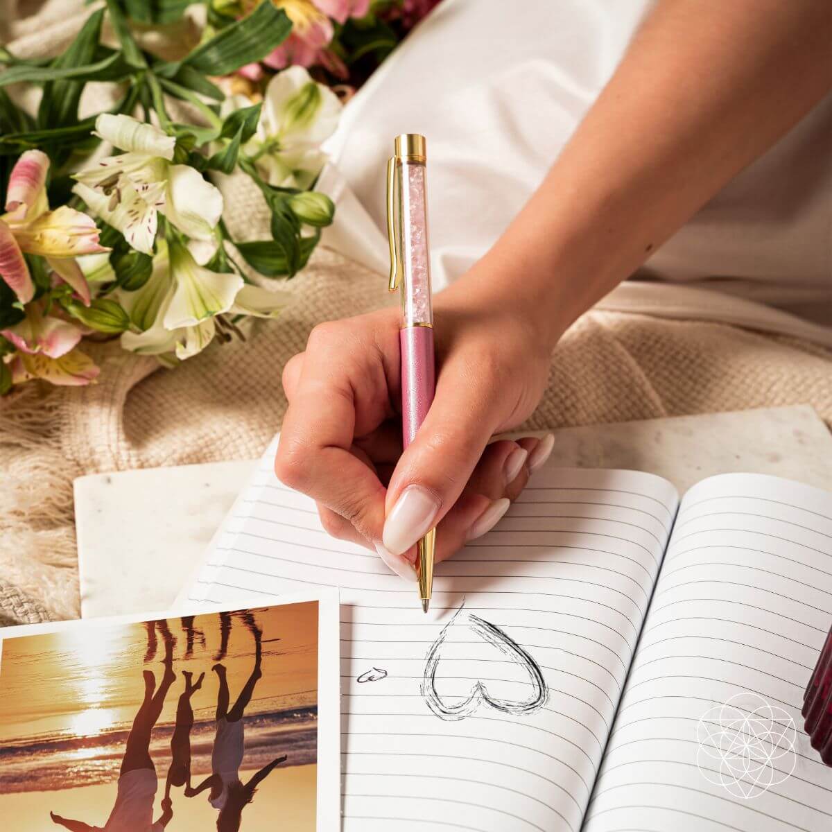 Hand drawing a heart in a lined notebook with a pink rose quartz crystal pen beside flowers.
