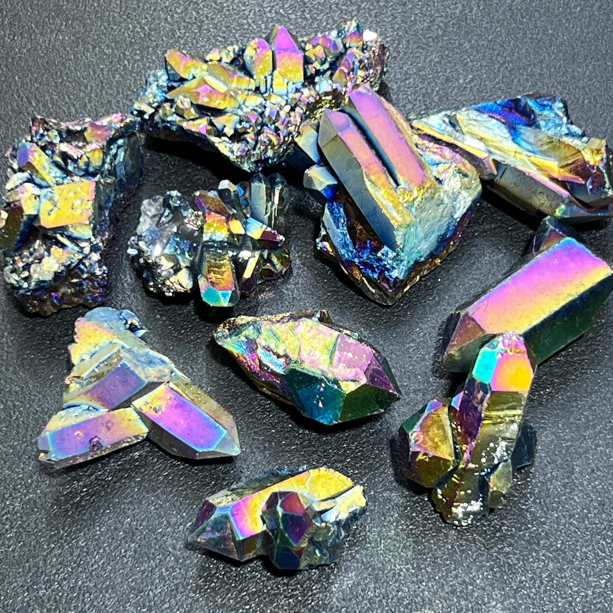 Small titanium aura quartz clusters and rough points with rainbow-colored crystal faces on a dark textured surface.
