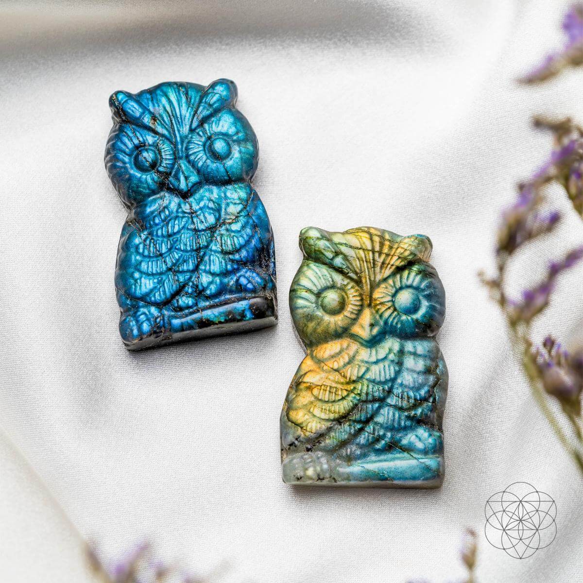 Blue and gold-flashing labradorite owl carvings lying on pale fabric beside small purple flowers.
