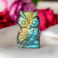 Carved labradorite owl with blue and gold flashes standing on pale fabric with pink flowers behind it.
