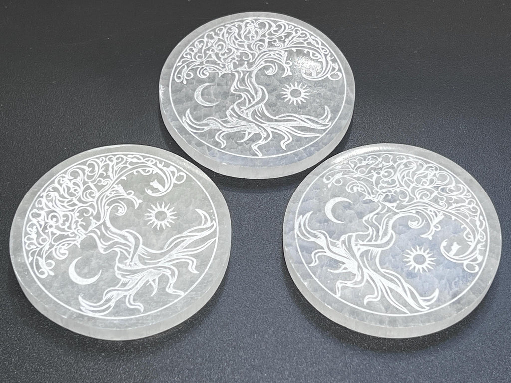 Three round white selenite plates engraved with Tree of Life, sun, and crescent moon designs on a dark surface.
