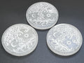 Three round white selenite plates engraved with Tree of Life, sun, and crescent moon designs on a dark surface.
