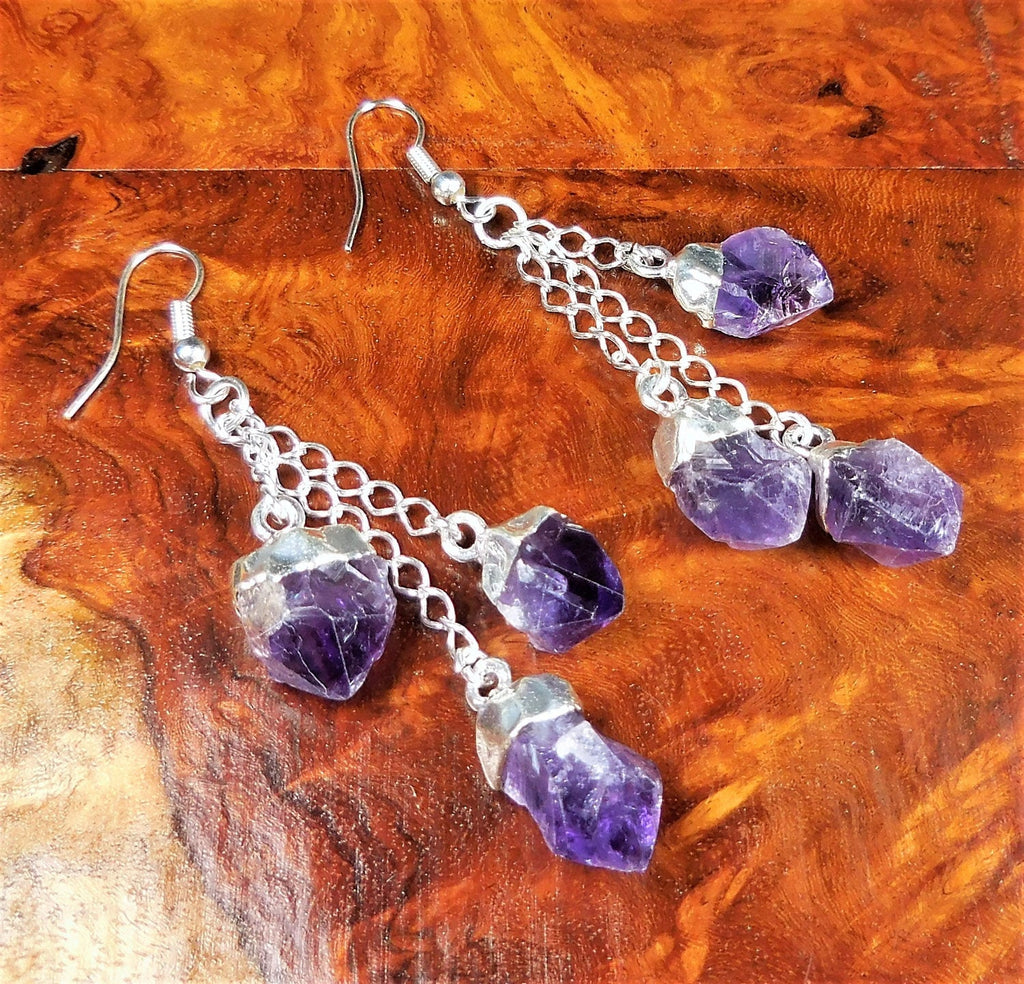 Pair of triple amethyst crystal earrings with silver-plated chains laid on reddish figured wood.