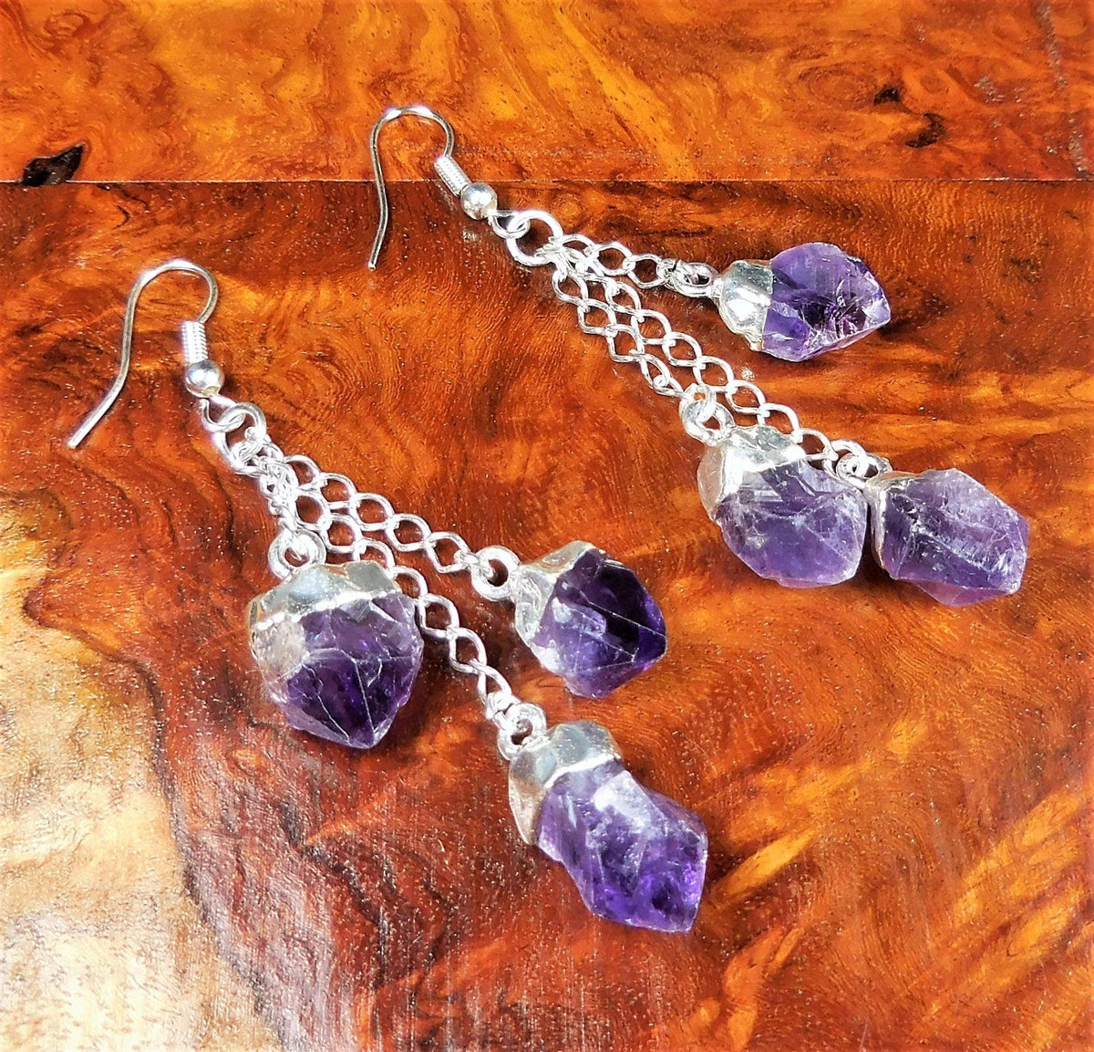 Pair of triple amethyst crystal earrings with silver-plated chains laid on reddish figured wood.