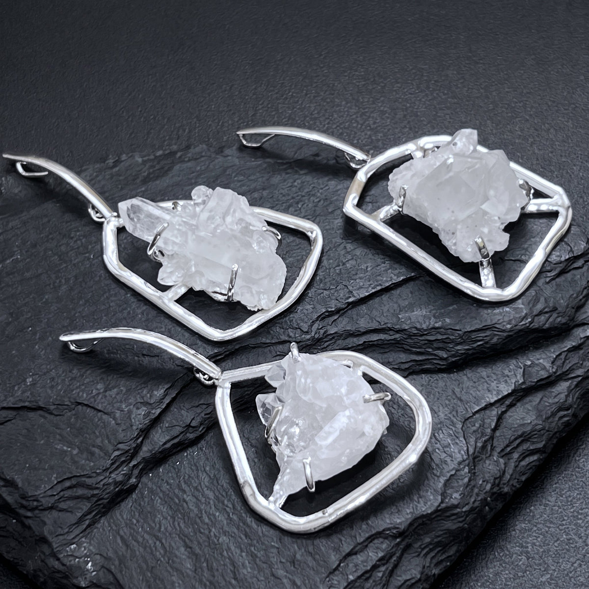 Three white quartz cluster pendants in silver-plated open frames with elongated bails arranged on dark slate.
