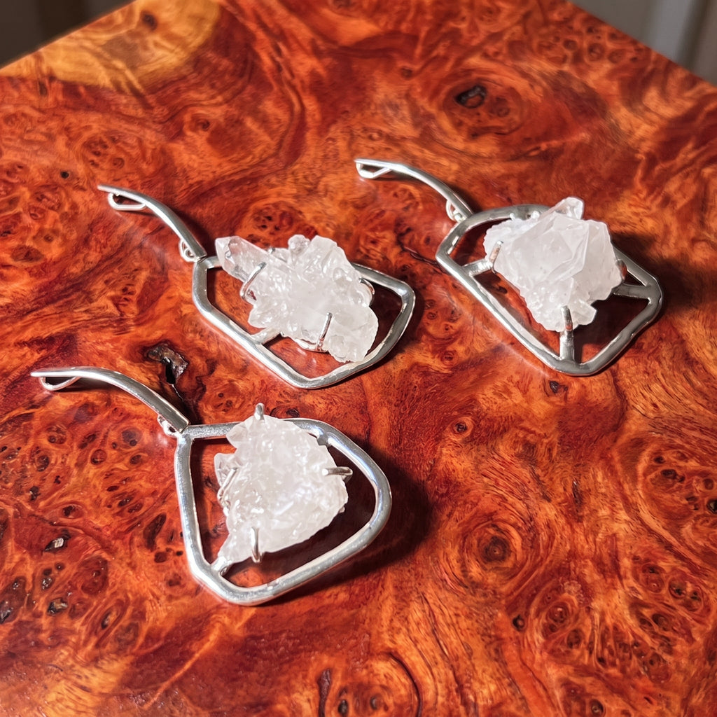 Three white quartz cluster pendants with silver-plated open settings and long bails on reddish patterned wood.
