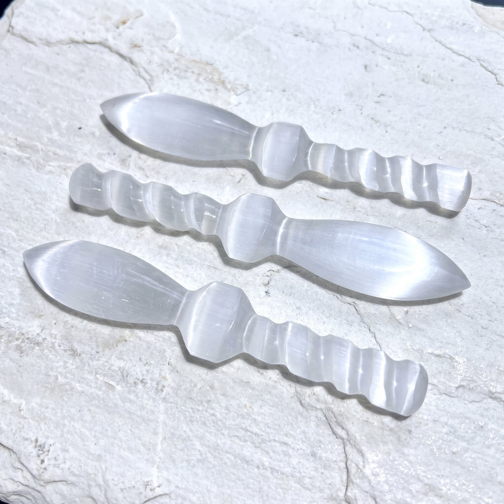 Three translucent white satin spar crystal daggers with spiral handles on pale stone in sunlight