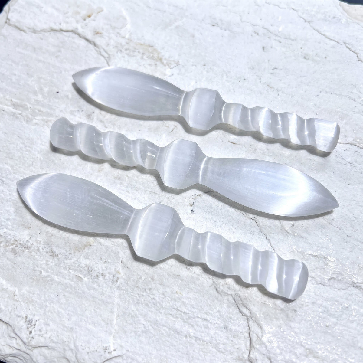 Three translucent white satin spar crystal daggers with spiral handles on pale stone in sunlight