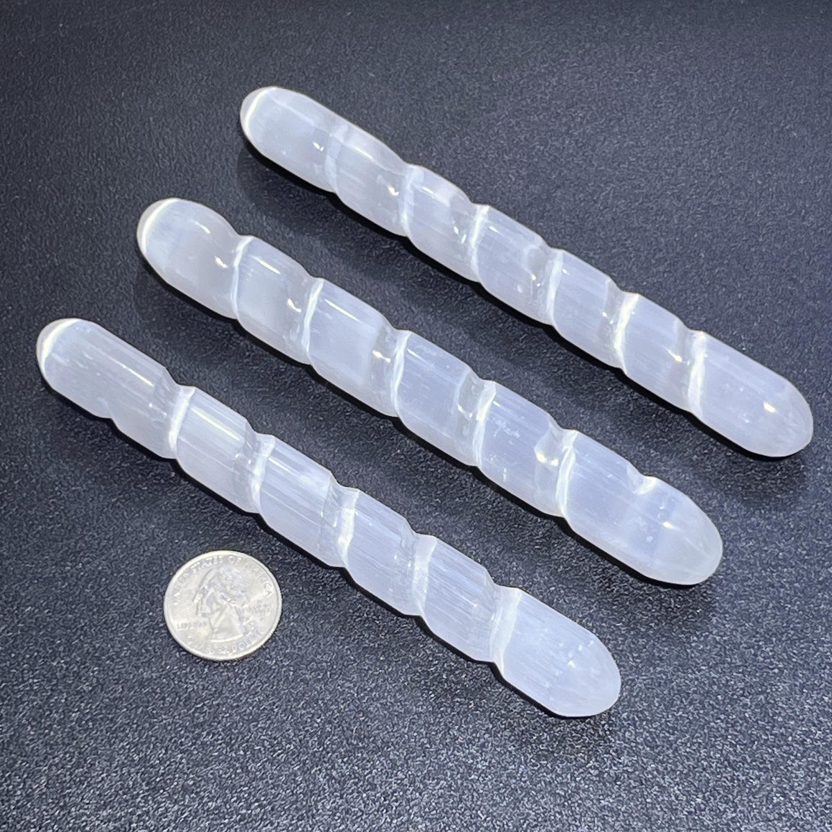 Three white satin spar selenite spiral wands with rounded ends beside a quarter on a dark surface.
