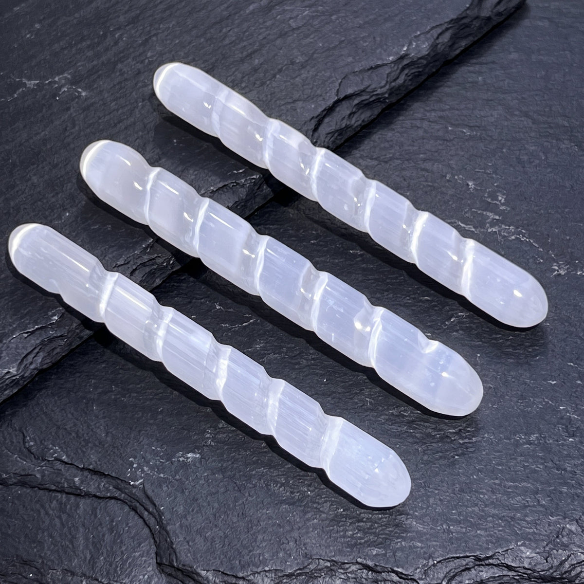 Three rounded white satin spar spiral wands arranged diagonally across layered dark slate.

