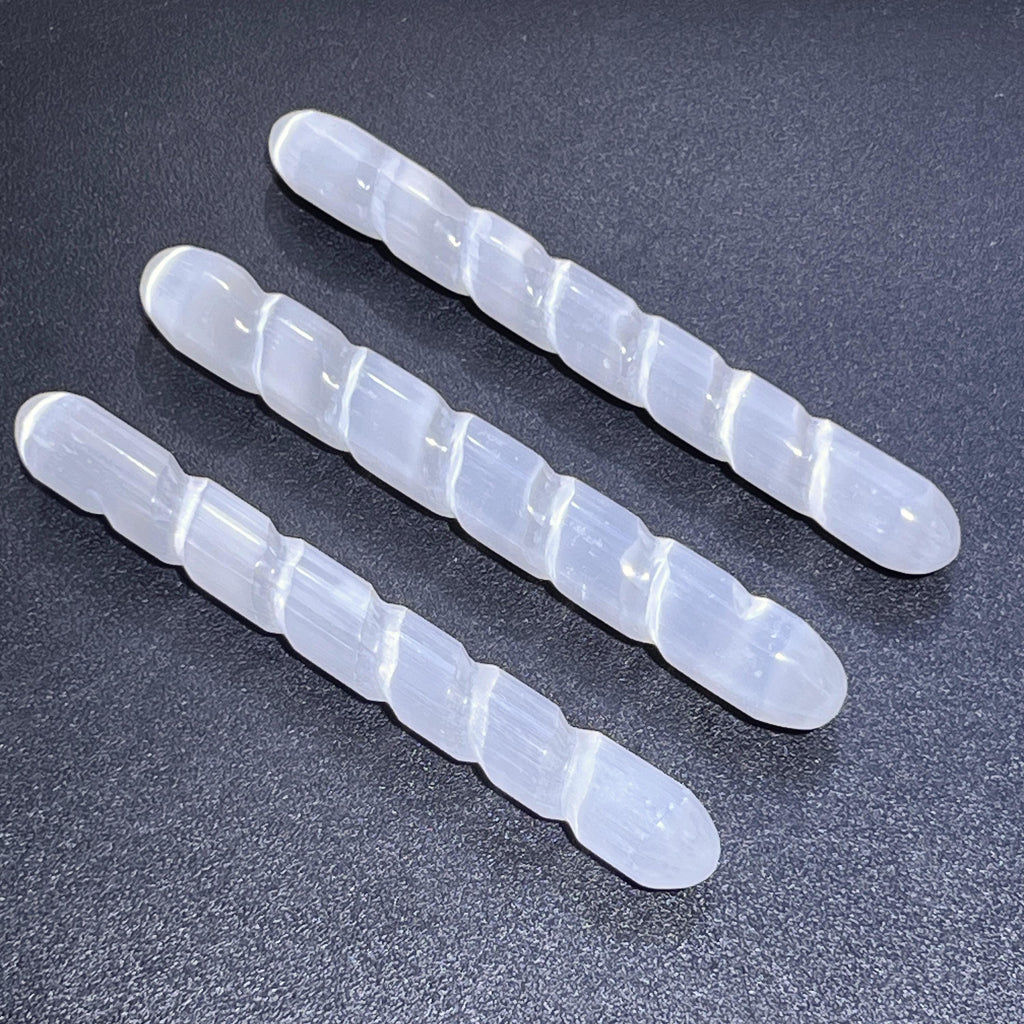 Three white selenite satin spar spiral wands showing rounded ends and light-catching carved grooves on a dark background.
